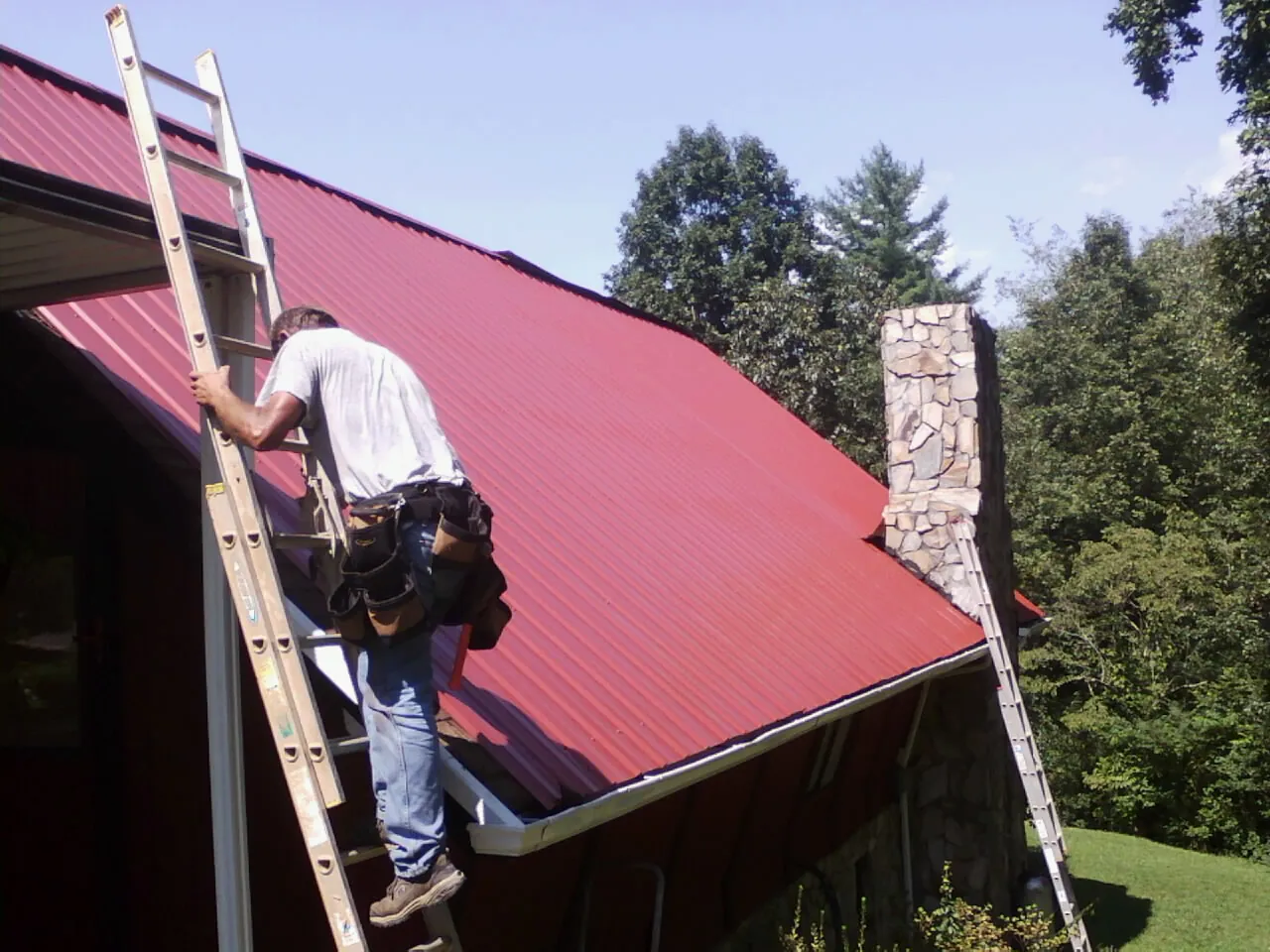 Roofer on a ladder beside a red metal roof and stone chimney — Soffit & Fascia Repair by Roofing Brothers in Elmore City